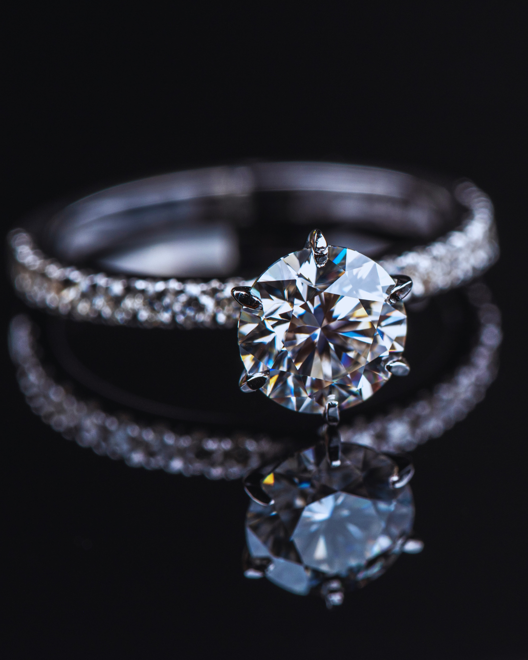 Primo piano di un anello solitario in oro bianco o platino con un grande diamante centrale rotondo incastonato e una fascia interamente tempestata di diamanti più piccoli, riflesso su una superficie scura