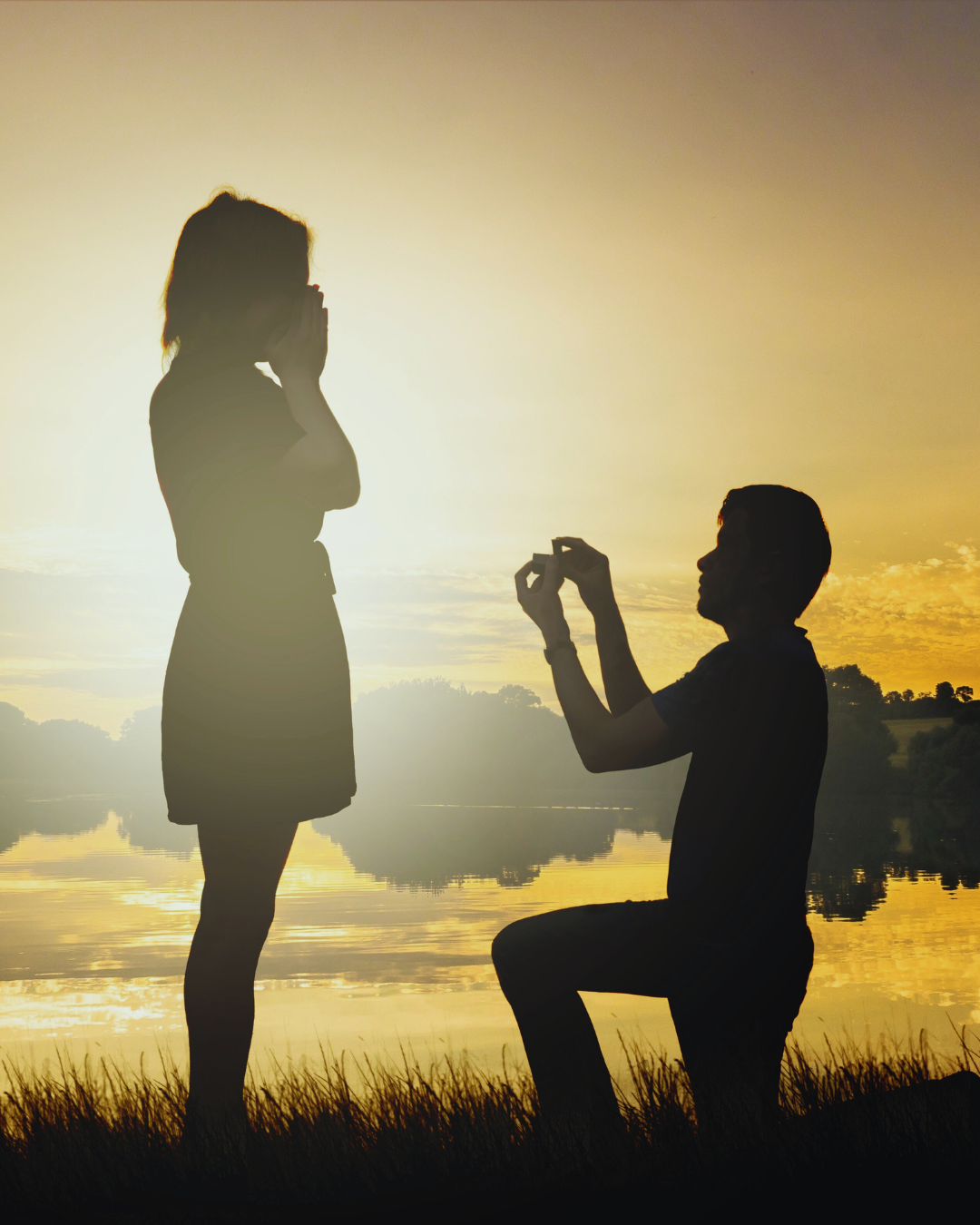 Silohuette di un uomo in ginocchio che fa la proposta di matrimonio a una donna, con un anello di fidanzamento, davanti a un lago al tramonto. Scena romantica e indimenticabile, simbolo di amore eterno da Atelier Diamanti