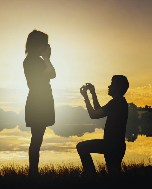 Silohuette di un uomo in ginocchio che fa la proposta di matrimonio a una donna, con un anello di fidanzamento, davanti a un lago al tramonto. Scena romantica e indimenticabile, simbolo di amore eterno da Atelier Diamanti