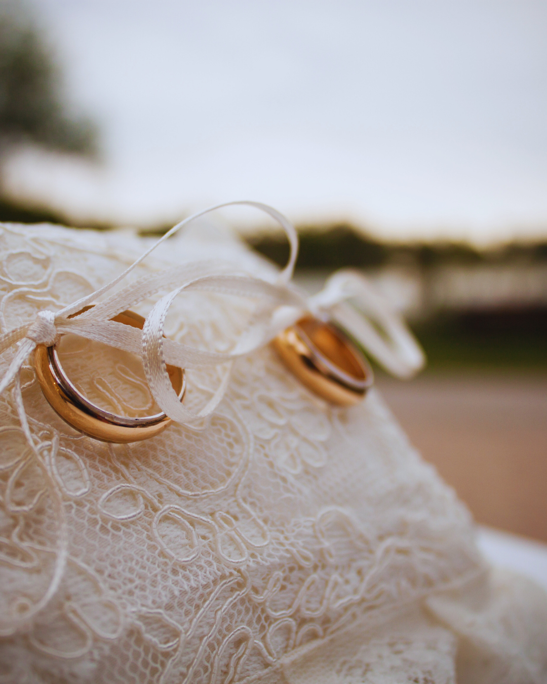 Due fedi nuziali in oro giallo lucido legate con un nastro bianco su un cuscino di pizzo bianco, pronte per una cerimonia nuziale. Simbolo di amore e unione di Atelier Diamanti