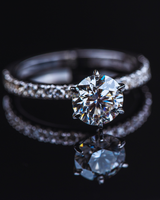 Primo piano di un anello solitario in oro bianco o platino con un grande diamante centrale rotondo incastonato e una fascia interamente tempestata di diamanti più piccoli, riflesso su una superficie scura
