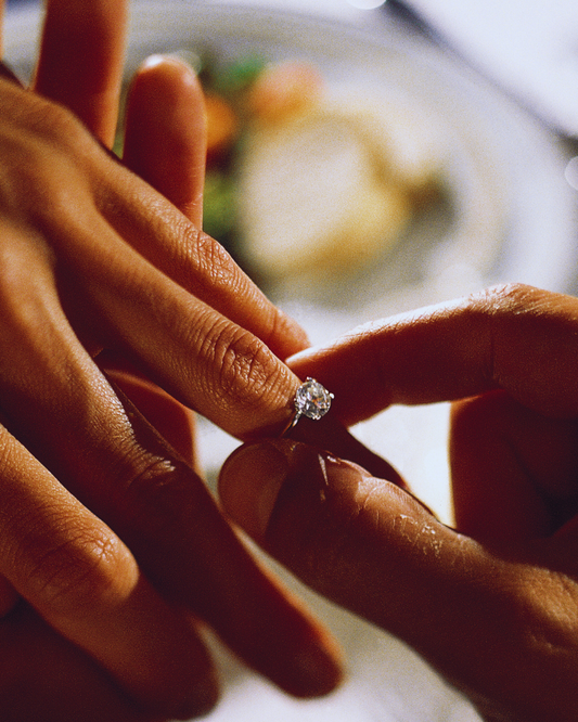 Mano di un uomo che mette un anello solitario con diamante al dito di una donna, in un momento intimo di proposta di fidanzamento o celebrazione. Gioiello d'amore eterno di Atelier Diamanti
